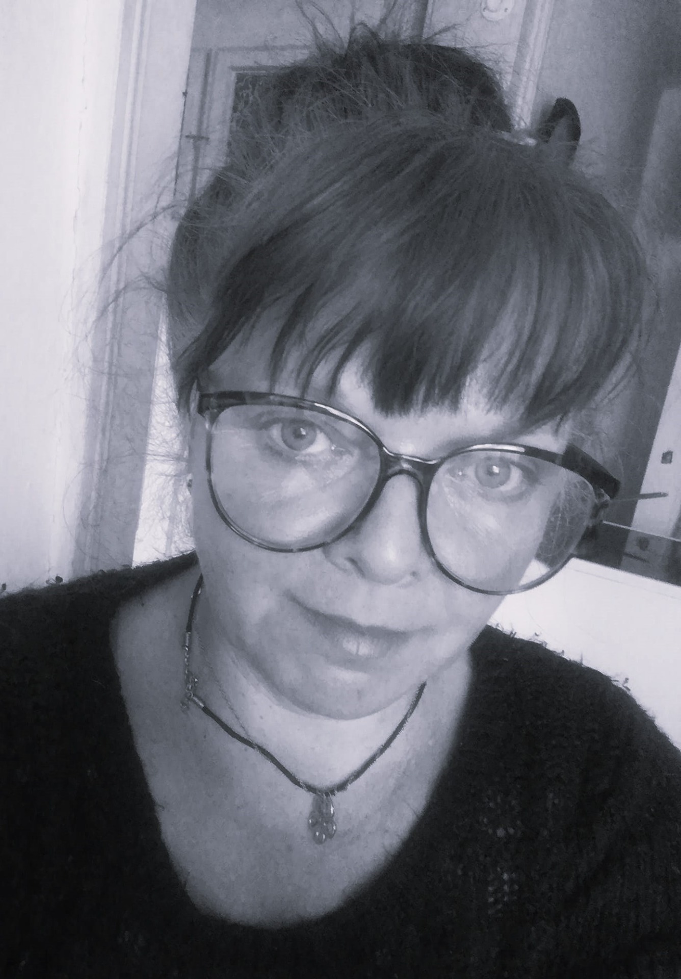 Černobílý selfie portrét ženy s brýlemi, krátkými vlasy a jemným úsměvem v interiéru. ano, uprav barevnost